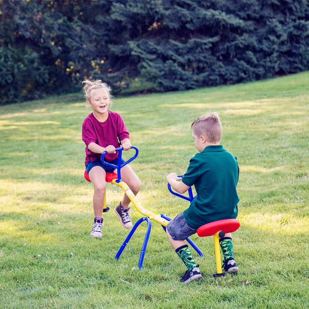 Two kids play on the red, yellow, and blue stationary Teeter Totter. 