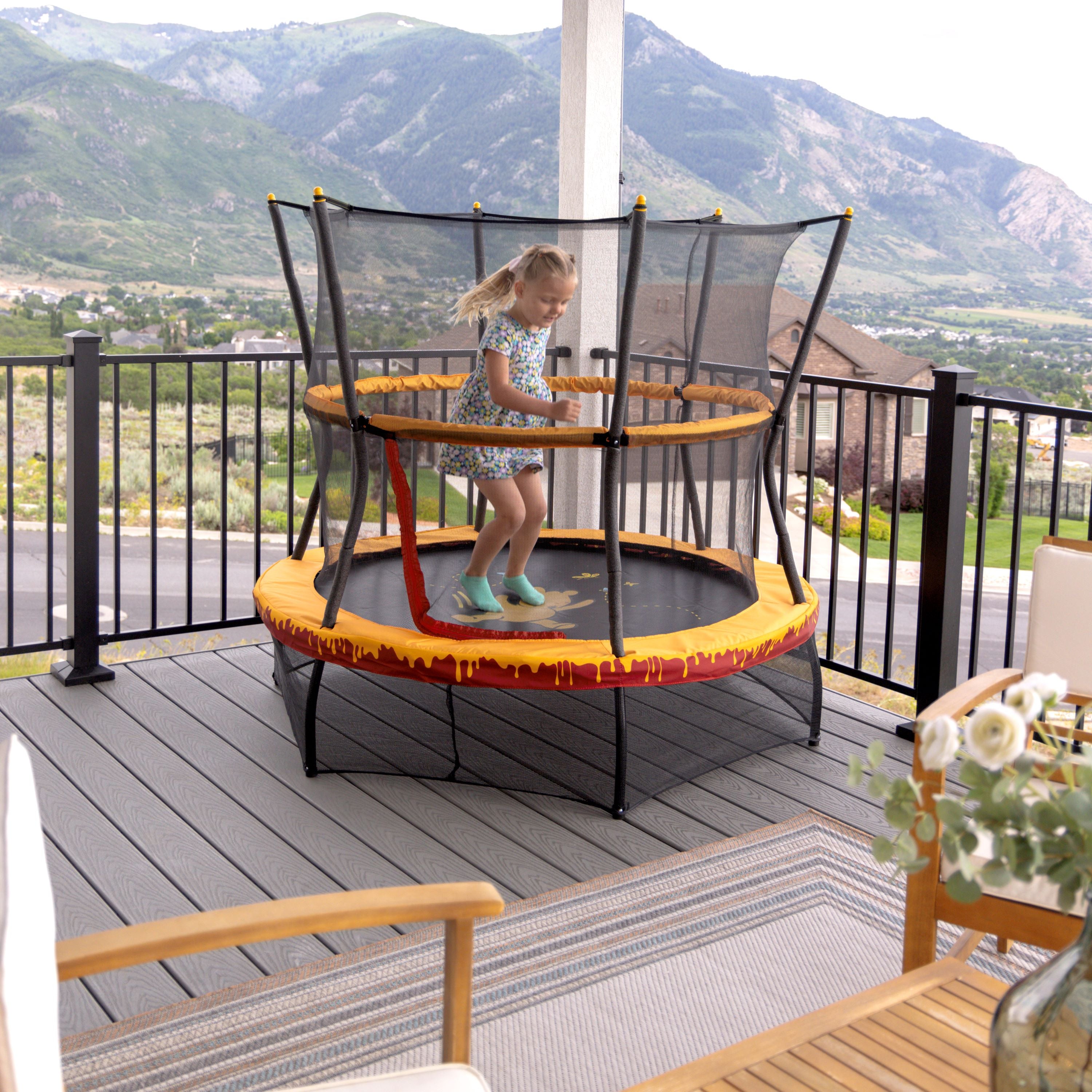 The 60” Winnie the Pooh toddler trampoline sits outdoors on the porch as a young girl jumps on it. 