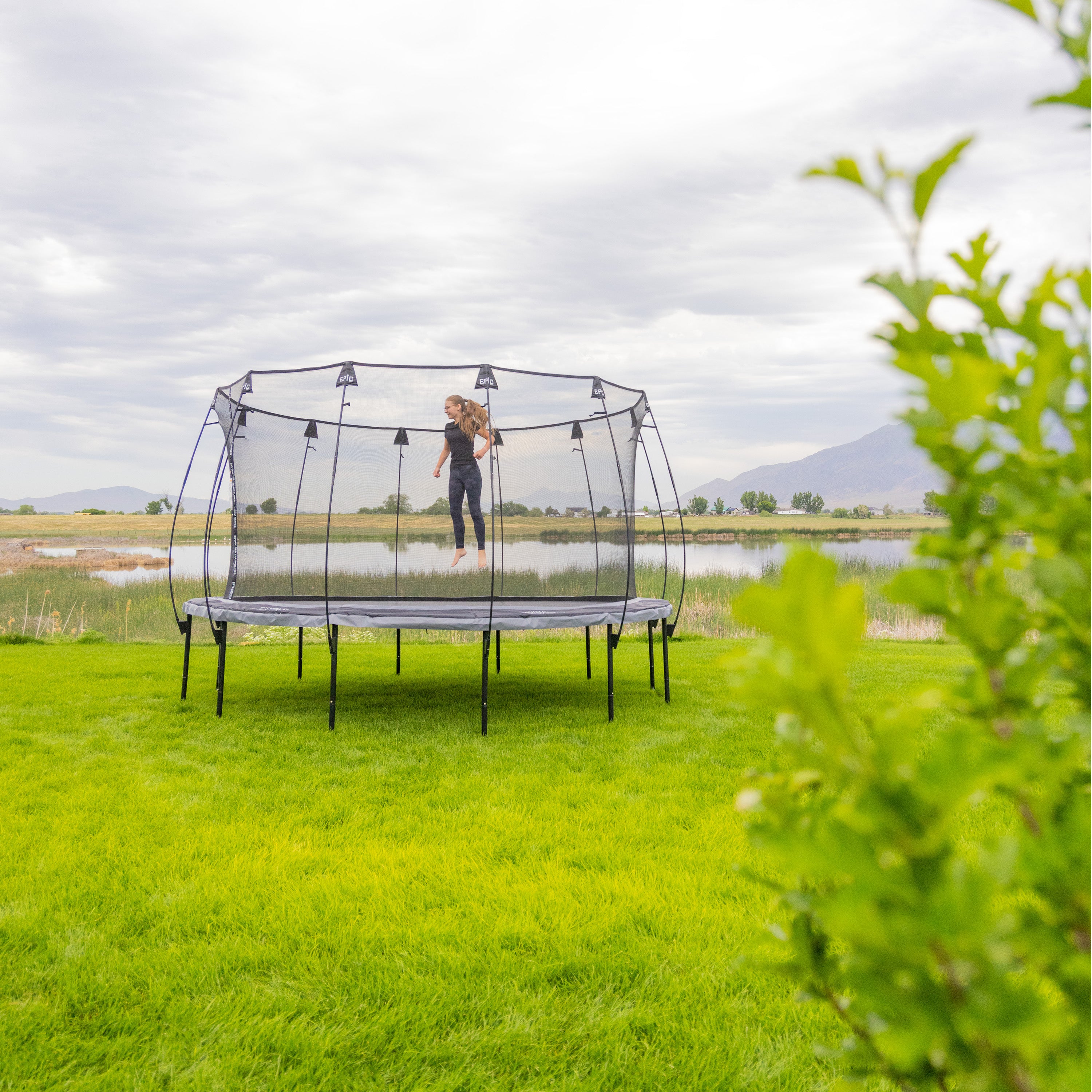 A girl dressed in all black jumps on the 14-foot Epic Series trampoline that is in a very green backyard. 