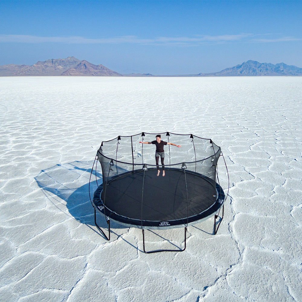 Teenage boy jumps with arms up on the 16-foot round Epic Series Trampoline with the Salt Flats in the background. 