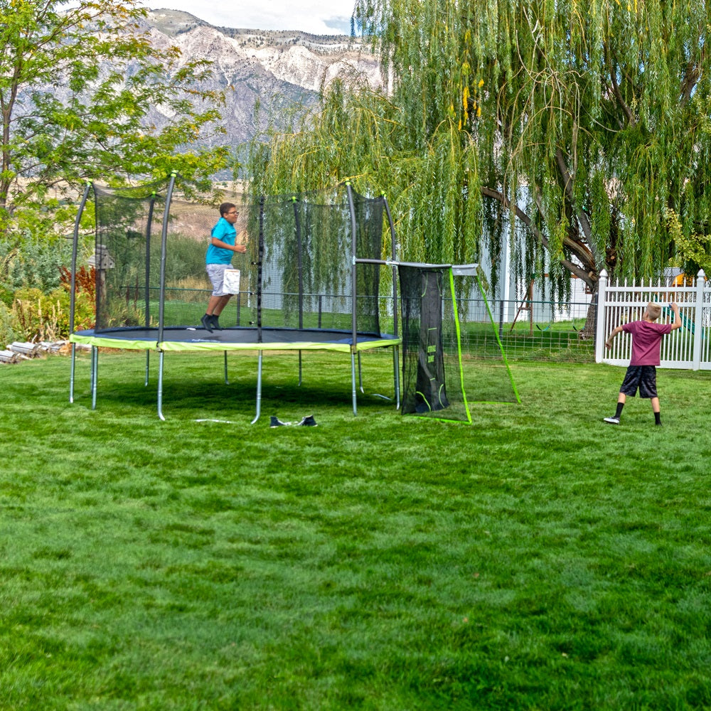 Boy pulling back arm to throw ball at the sports net while another boy jumps on the trampoline. 