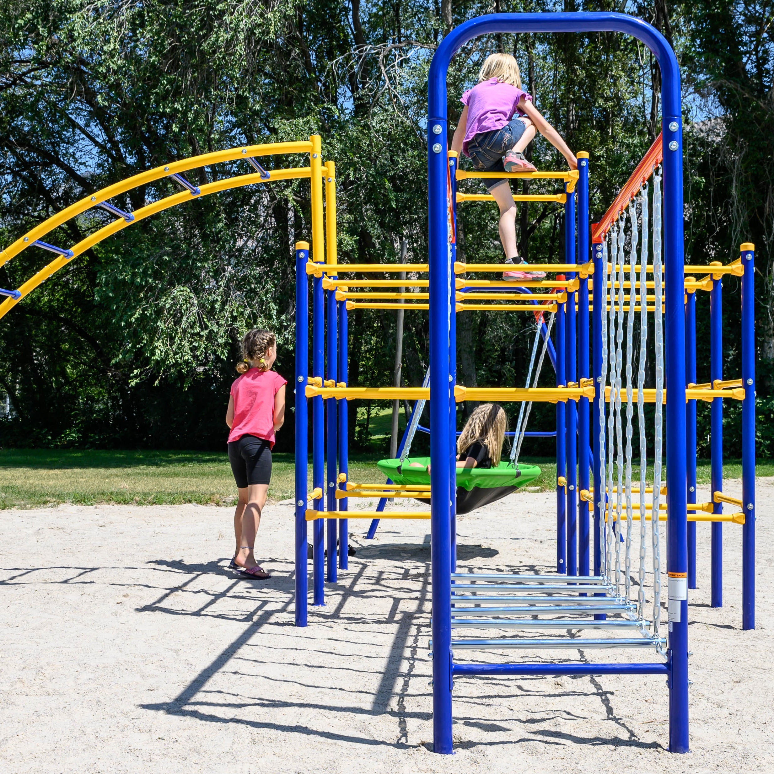 Three young girls climb on the playset that includes the Saucer Swing, Modular Jungle Gym, Hanging Bridge, and Arched Ladder. 