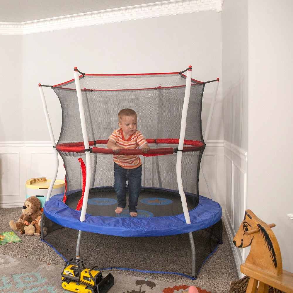 Little boy stands on mini trampoline while holding red padded handlebar. Toys are scattered on the floor around him.