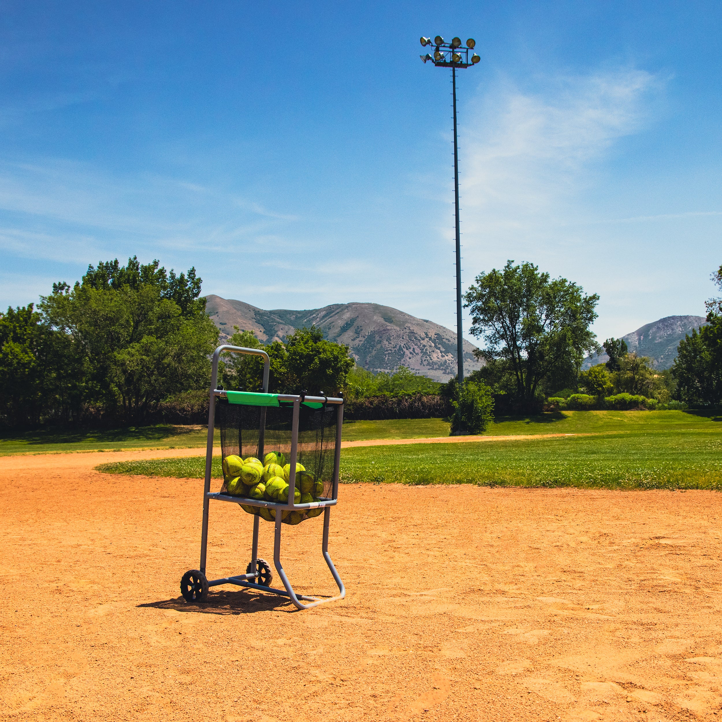 The Baseball and Softball Cart with Wheels holds softballs inside while sitting on a baseball field. 
