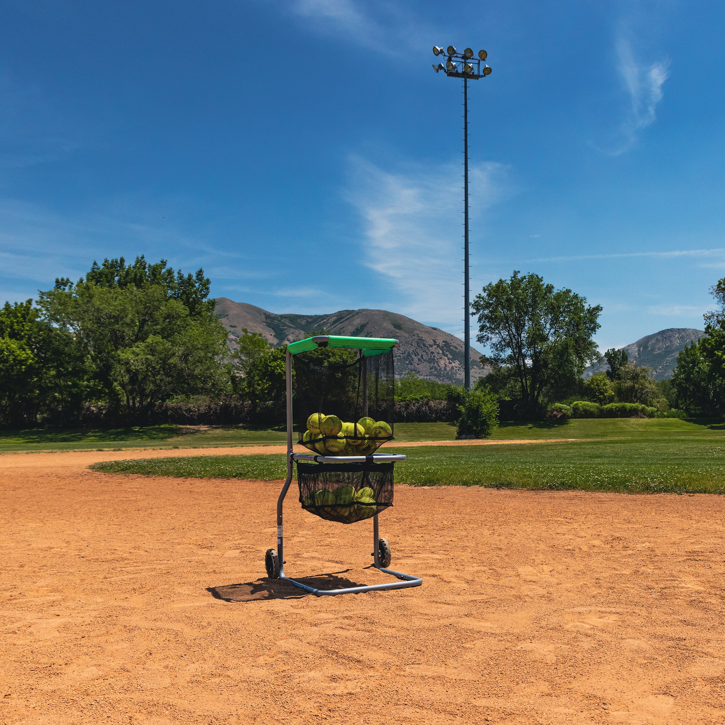 The Multi-Sport ball cart sits on a baseball field with more than a dozen softballs in the two baskets. 