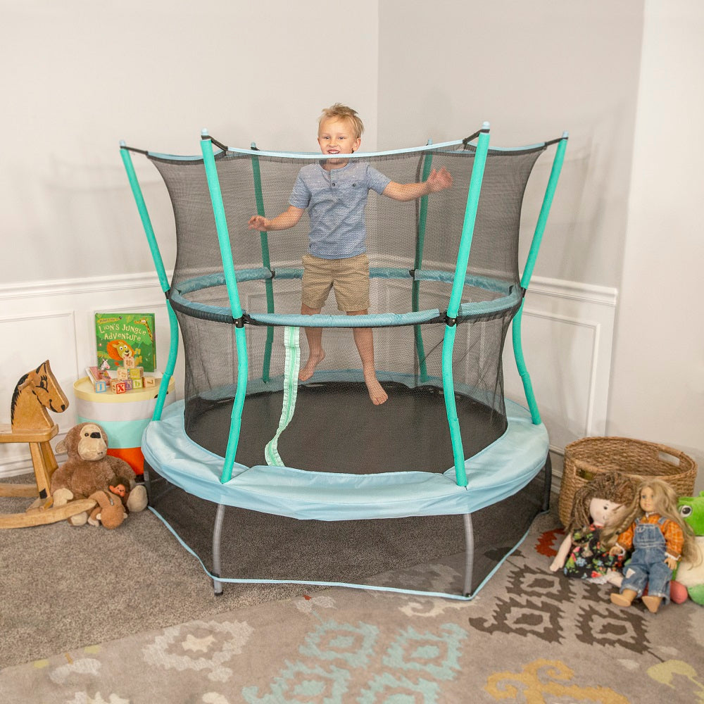 Young toddler jumps on 60-inch indoor kids trampoline with blue frame pad and seafoam green enclosure poles.