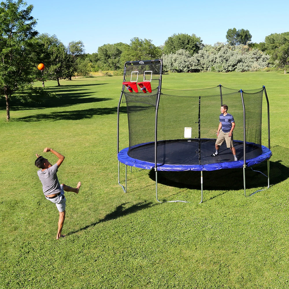 One boy jumps on the trampoline while another boy tries to shoot a basket from outside the trampoline. 