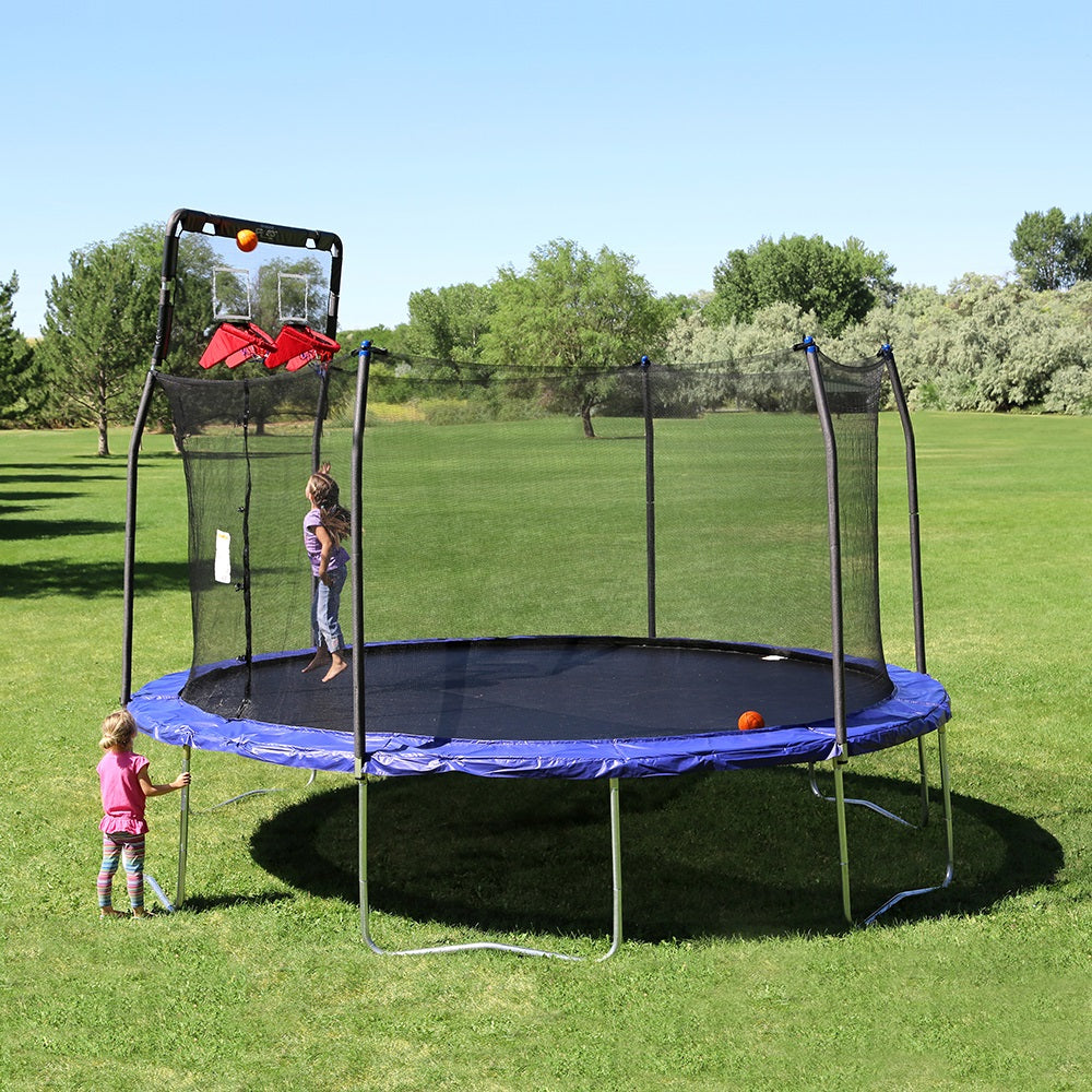 A little girl shoots a basketball towards the double basketball hoop while another girl watches from outside the trampoline. 