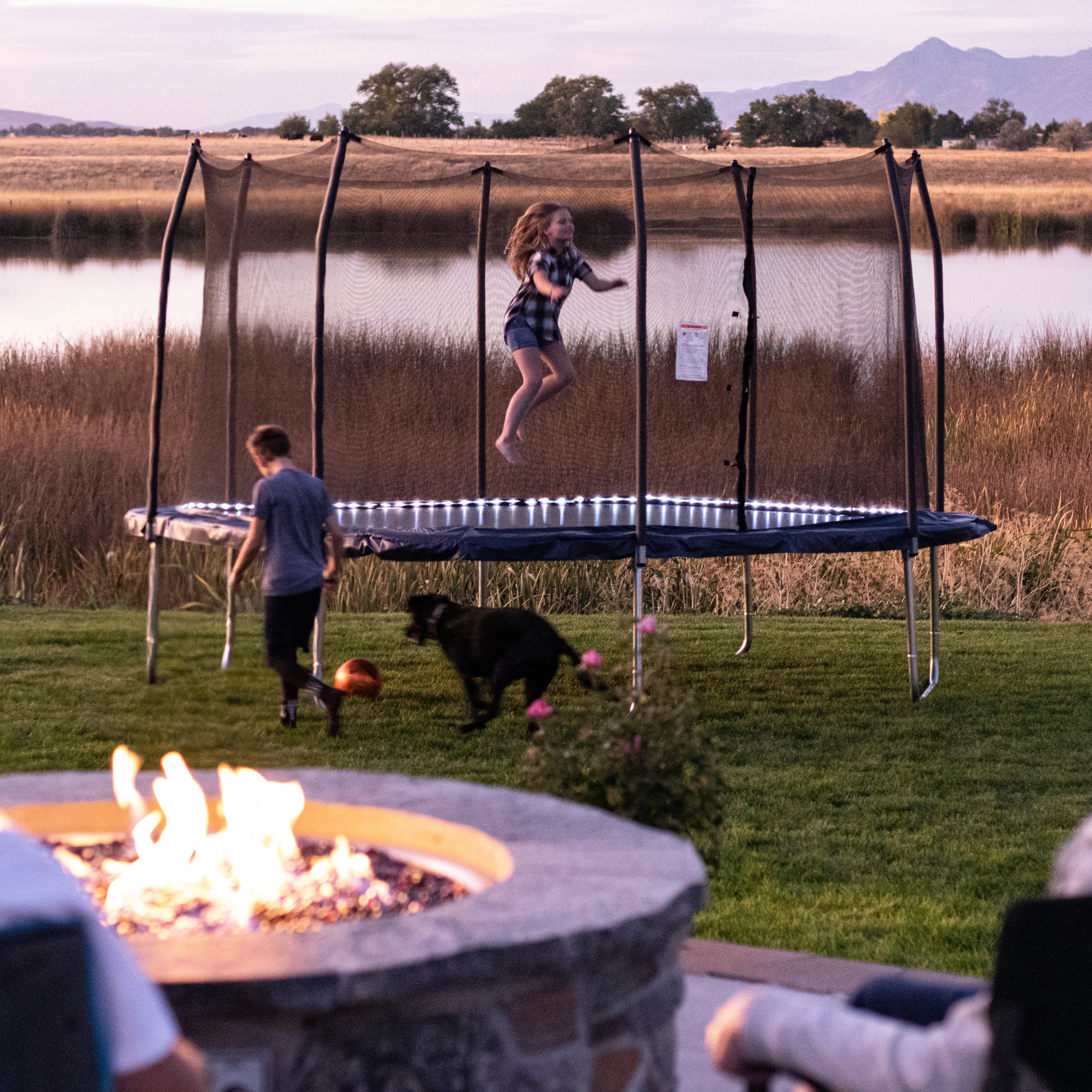 Girl jumps on 13' square trampoline with lighted spring pad. Boy and dog play in grass, and parents watch while sitting by fire. 