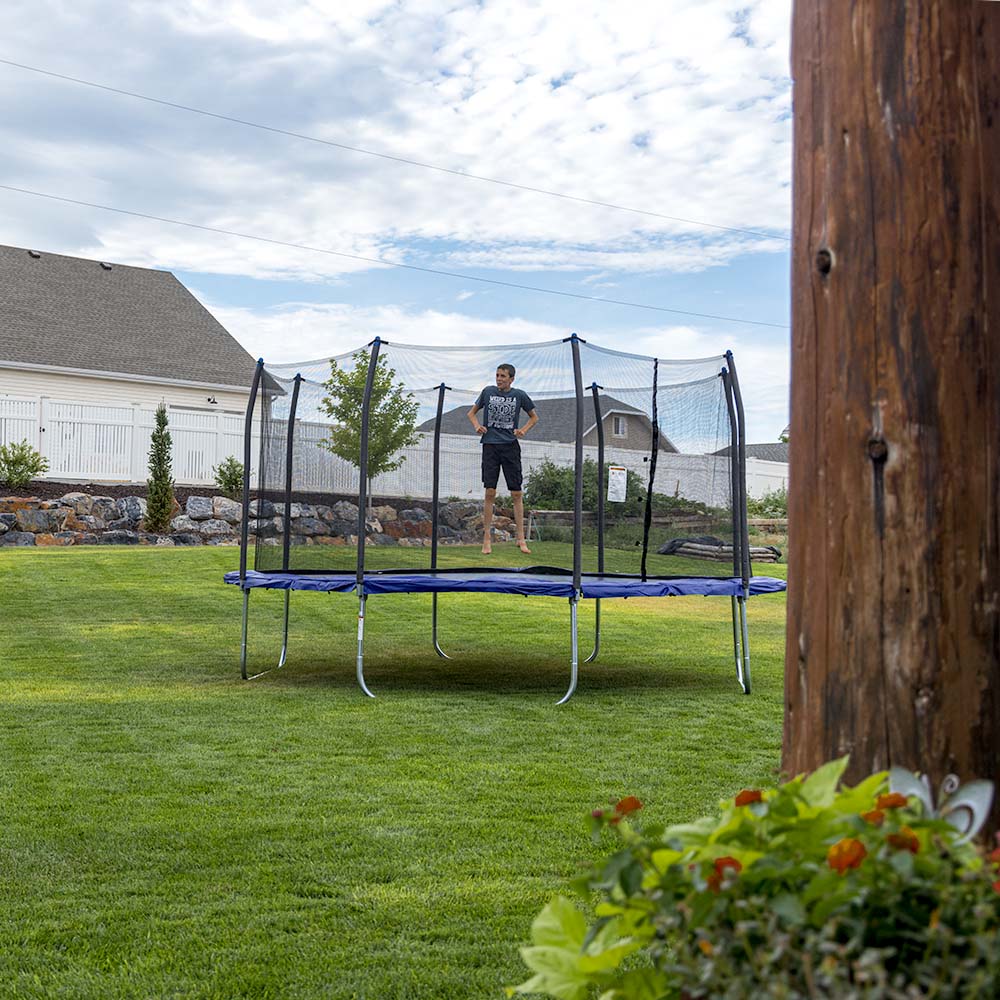 Young boy bounds on fourteen-foot square trampoline with blue spring pad and blue pole caps.