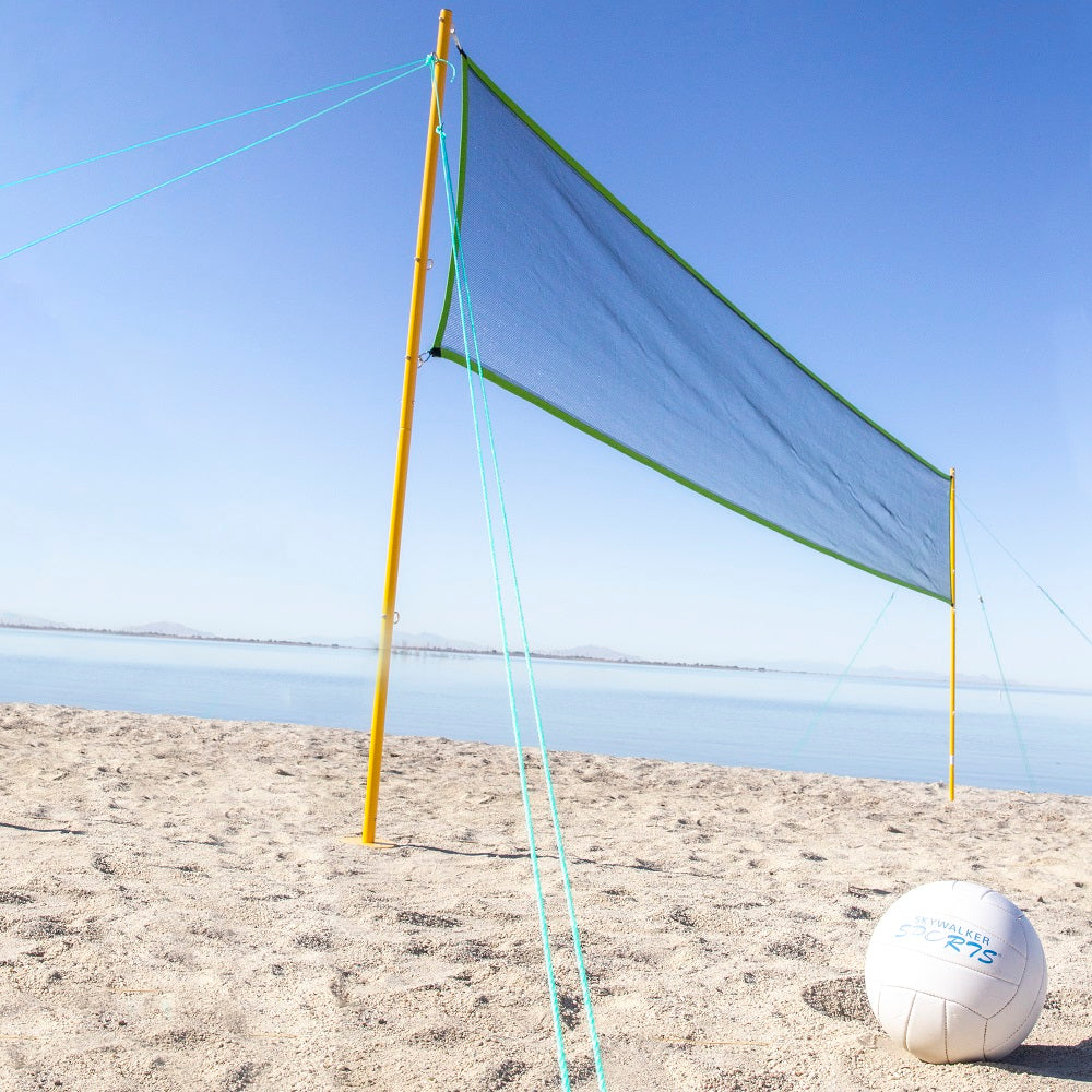 The volleyball net is staked into a sandy beach. The volleyball sits in the sand in front of it. 