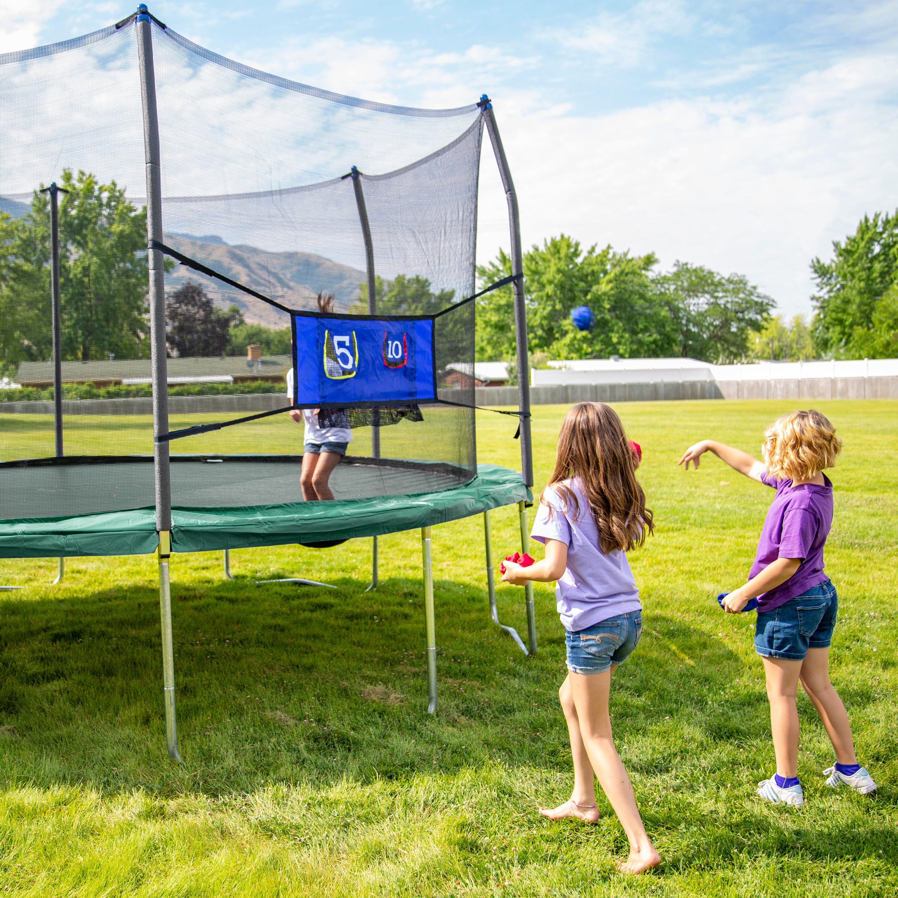 Two girls throw bean bags at the Double Toss game while another girl jumps on the trampoline. 